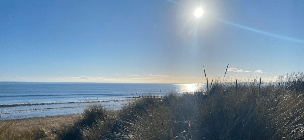Looking out to sea, at Dawlish Warren, with the dunes in the foreground, a very blue sky, flat sea and a bright sun and sparkles. 